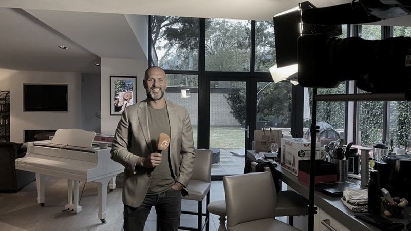Een televisiecamera op statief in een woonkamer, Rico Briedjal ernaast